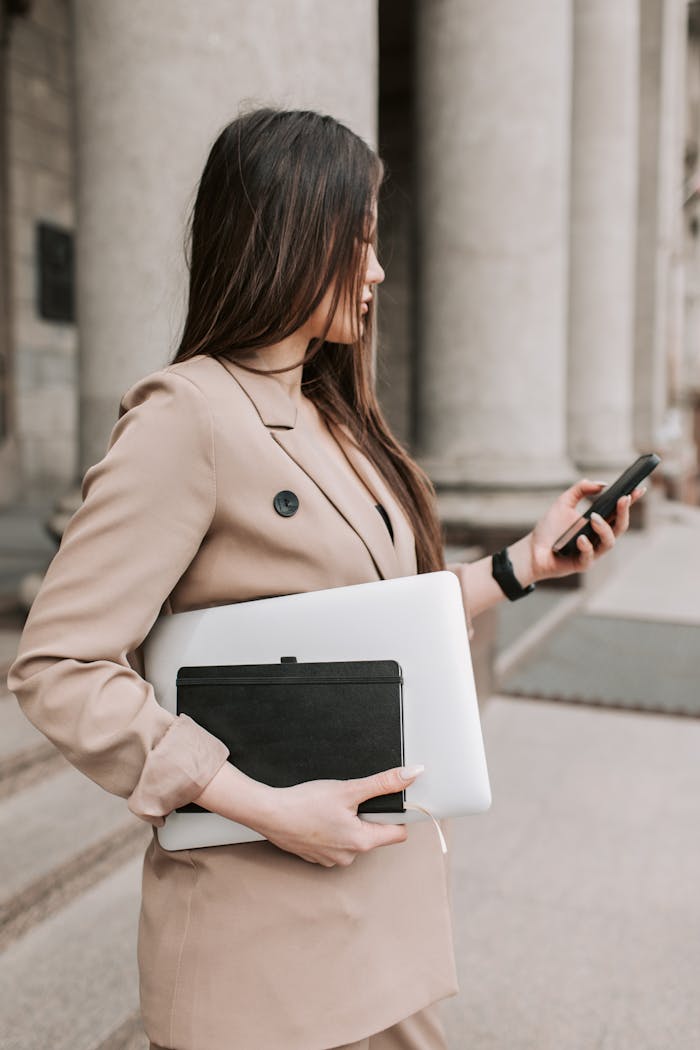 why-choose-us Confident young woman checking her phone while holding a laptop and notebook outside a building.